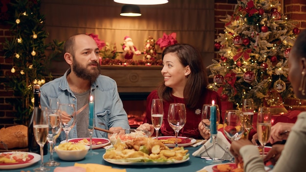 Festive couple celebrating Christmas at evening dinner table with close family members. Positive people sitting in living room to celebrate authentic traditional winter holiday with loved ones.