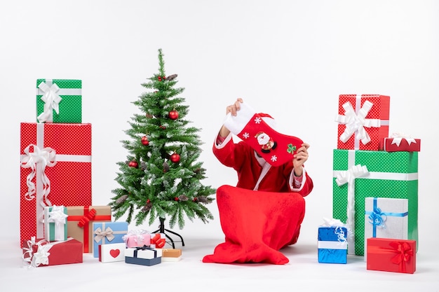 Free Photo festive holiday mood with santa claus sitting on the ground and closing his face with christmas sock near gifts and decorated xsmas tree on white background