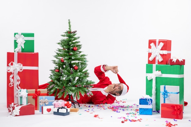 Free Photo festive holiday mood with young surprised santa claus lying behind christmas tree near gifts on white background