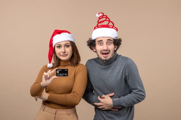 Free photo festive mood with cool couple wearing red santa claus hats girl showing bank card on gray