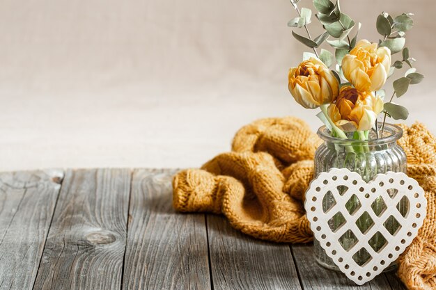 Festive still life for valentine's day with flowers in a vase, knitted element and decorative heart on a wooden surface close up.