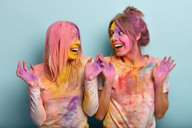 Free photo festivity and colored holiday concept. optimistic european women raise hands and discuss something happily, have fun together, play with colors, express good emotions. holi festival in india