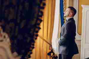 Free photo the fiance in a black suit and a beard stands in a room near the window