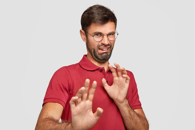 Free photo fie! dissatisfied bearded male pushes hands away, has disgusting facial expression, notices something detestable, frowns face with aversion, wears red t-shirt, isolated over white wall