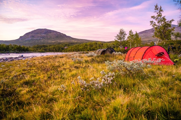 Field with tents surrounded by hills covered in greenery under a cloudy sky during the sunset