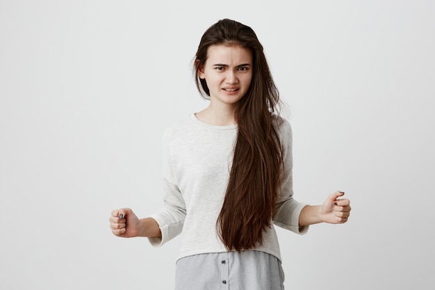 Free photo fierce and angry brunette female model in casual clothes holding fists in irritation, frowning face, clenching white teeth having dassatisfied expression. negative emotions, feelings and reaction