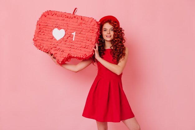 Fiery red curly lady in hat and dress is looking at camera and holding like sign on isolated space.