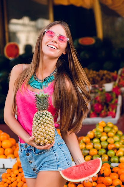 Free photo filled with joy summer girl having fun on tropical fruits market. she holds ananas, slice of watermelon and smiling