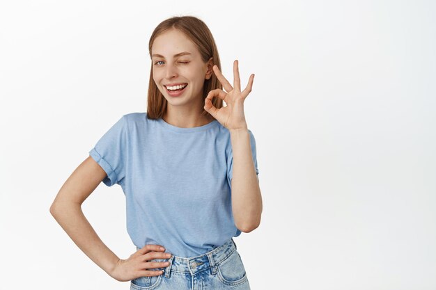 Fine, no problem. Smiling blond woman assure you, show okay OK sign and winking satisfied, praise excellent work, good job, standing in t-shirt over white background.