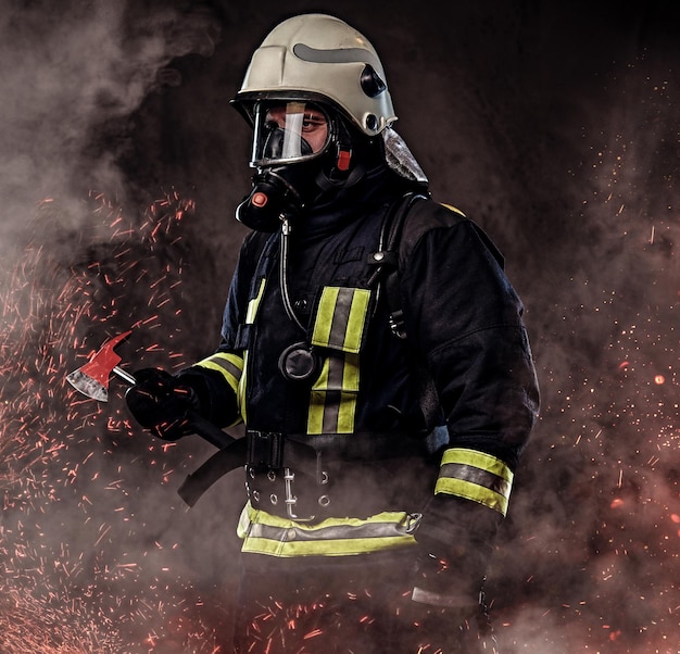 Free photo a firefighter dressed in a uniform and an oxygen mask holds a red axe standing in fire sparks and smoke over a dark background.