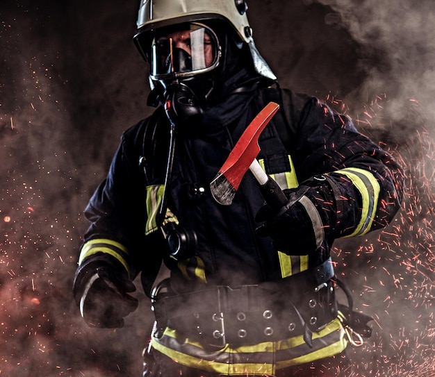 A firefighter dressed in a uniform and an oxygen mask holds a red axe standing in fire sparks and smoke over a dark background.