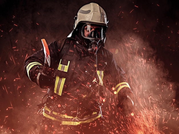 Free photo a firefighter dressed in a uniform and an oxygen mask holds a red axe standing in fire sparks and smoke over a dark background.