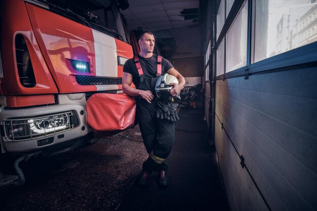 Fireman holding a jacket and protective helmet in a garage of a fire department, leaning on a fire engine and looking outside