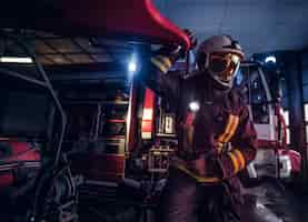 Free photo fireman wearing protective uniform standing next to a fire engine in a garage of a fire department