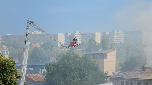 Firemen on platform truck going on roof of house on fire. View of firefighters trying to extinguish fire from burning building in flames and fumes. Men stopping smog and smoke from house.