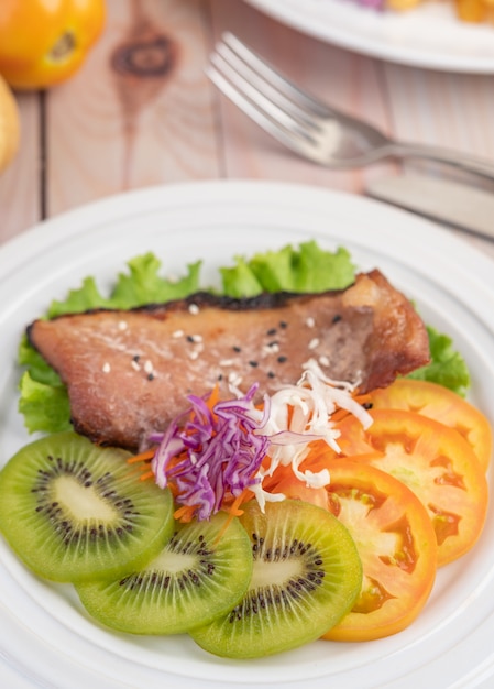 Free Photo fish steak with french fries, kiwi, lettuce, carrots, tomatoes, and cabbage in a white dish.