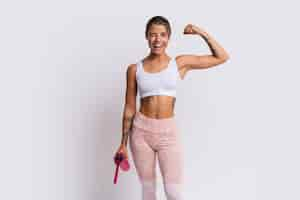 Free photo fit blond woman with perfect smile in stylish sportswear looking at camera and holding bottle of water over white wall. demonstrate muscles.