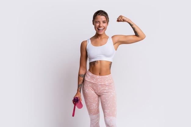 Fit blond woman with perfect smile in stylish sportswear looking at camera and holding bottle of water over white wall. Demonstrate muscles.