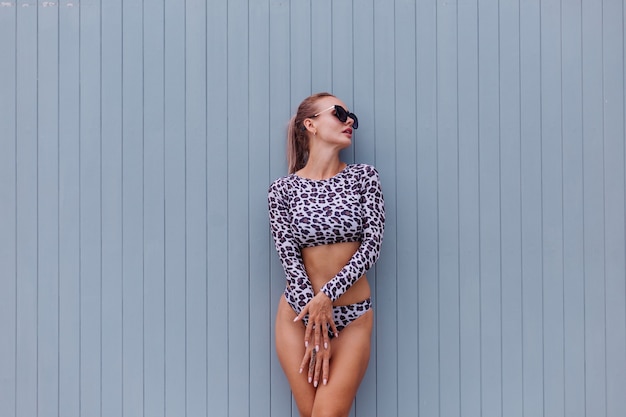 Fit caucasian woman in leopard swimsuit and sunglasses on gray wall