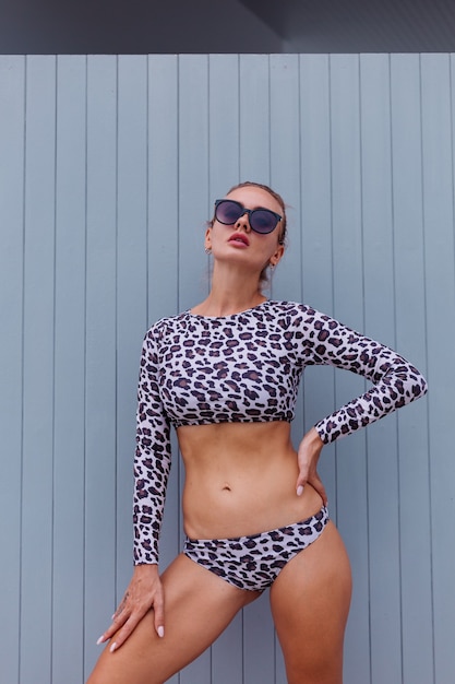 Free photo fit caucasian woman in leopard swimsuit and sunglasses on grey wall