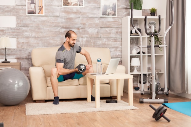 Free Photo fit man on sofa training biceps with dumbbell watching online workout in time of self isolation.