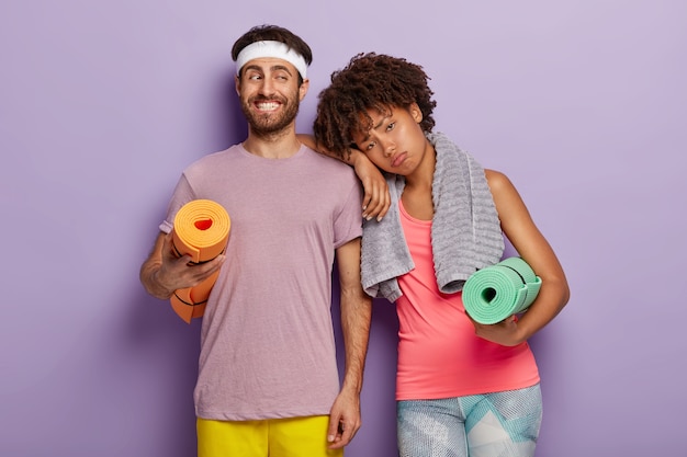 Free photo fit positive man with stubble carries rolled up karemat, tired woman leans at husbands shoulder, being tired after exhausting workout, has towel on neck, spend free time together in gym. sport