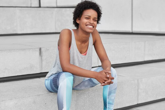 Fit satisfied dark skinned woman with Afro haircut, wears sportsclothes, keeps hands together, has glad expression, sits at stairs, feels refreshed, full of energy after cardio training. Sport concept