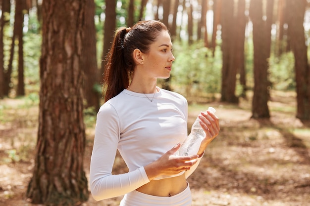 Fitness beautiful woman with dark hair and ponytail holding bottle of water and looking away, posing after exercising in forest