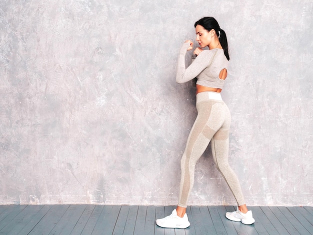 Fitness confident woman in sports clothing Sexy young beautiful model stands in a rack Female posing near grey wall in studio She practice power punches boxing during workout process