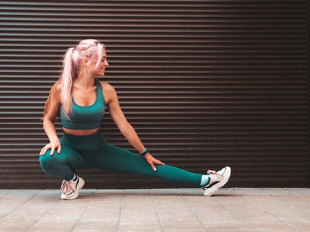 Free photo fitness smiling woman in green sports clothing with pink hair young beautiful model with perfect bodyfemale posing in the street near roller shutter wall stretching out before training