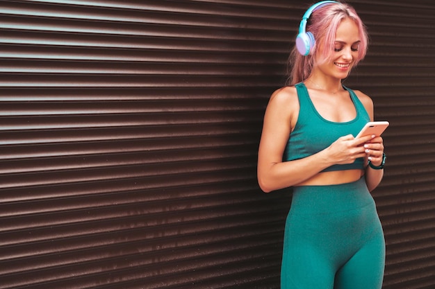 Free photo fitness smiling woman in green sports clothing with pink hair young beautiful model with perfect bodyfemale in the street near roller shutter walllistening music in wireless headphonesusing apps