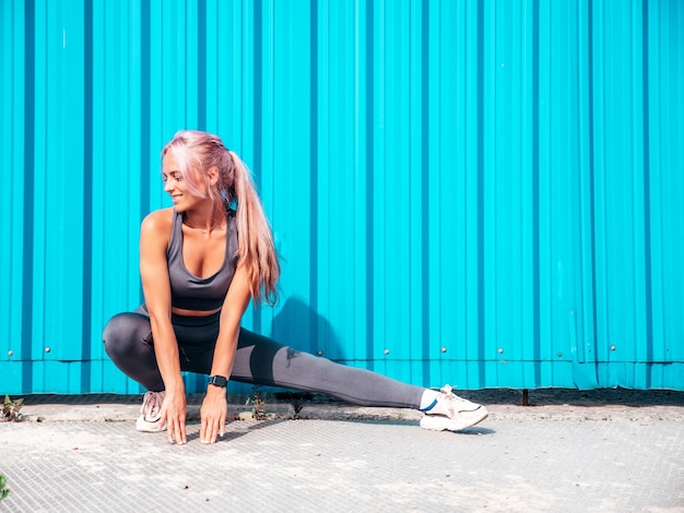 Free photo fitness smiling woman in grey sports clothing with pink hair young beautiful model with perfect bodyfemale posing in the street near blue wallcheerful and happy stretching out before training