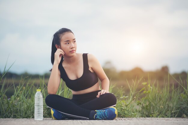  Fitness woman runner sit down relaxing with water bottle after training outside in park