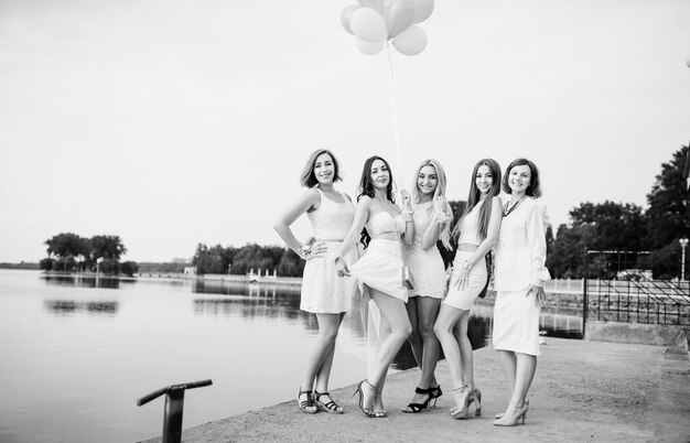 Five girls with balloons at hand weared on white dresses on hen party against pier on lake