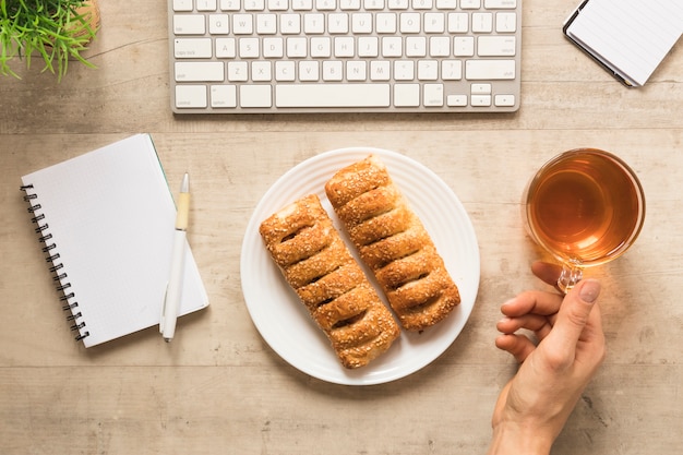 Flat lay hand holding tea cup near plate of pastry with notebook