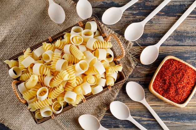 Flat lay macaroni pasta in tray with spoons, red spice on sackcloth and wooden background. horizontal