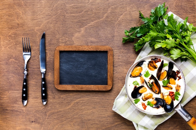 Flat-lay mussels in white sauce on tablecloth with blackboard