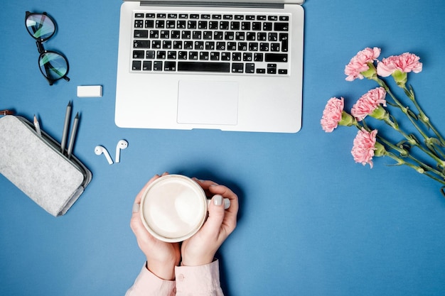 Flat lay woman office desk Female hands with coffee and laptop pink carnation stationery on blue table Top view