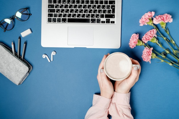 Free photo flat lay woman office desk woman hands with coffee and laptop pink carnation stationery on blue table top view