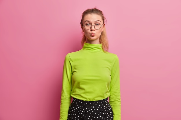 Free photo flirty girl keeps folds lips in glamour way, sees handsome guy, flirts, wants to kiss someone, wears green turtleneck, round big spectacles