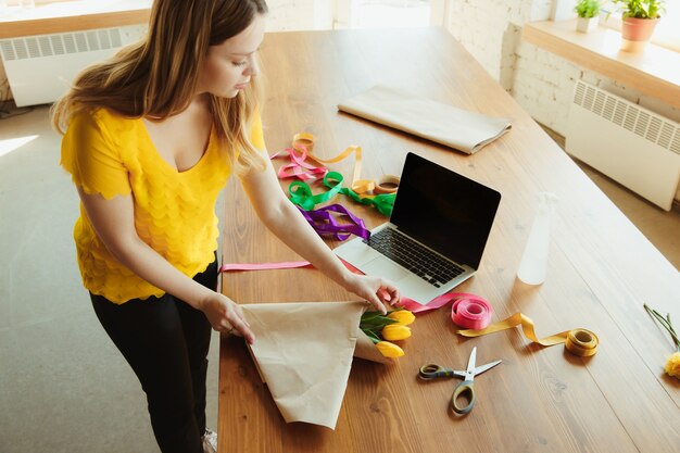 Florist at work: woman shows how to make bouquet with tulips. Young caucasian woman gives online workshop of doing gift, present for celebration. Working at home while isolated, quarantined concept.