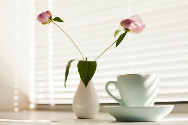 Flower vas and coffee cup with saucer near window blind