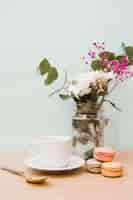 Free photo flowers in jar with cup; spoon and macaroons on wooden desk against wall