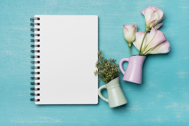 Flowers in two ceramic vases with blank spiral notebook on blue wooden background