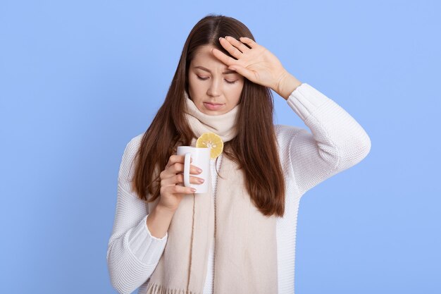 Flu treatment. Portrait of ill young woman wrapped in scarf drinking hot tea beverage, has seasonal influenza, viral infection, posing isolated on blue wall.