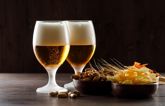 Foamed beer with pistachio, wheat ears, chips in goblet glasses on wooden table, side view.