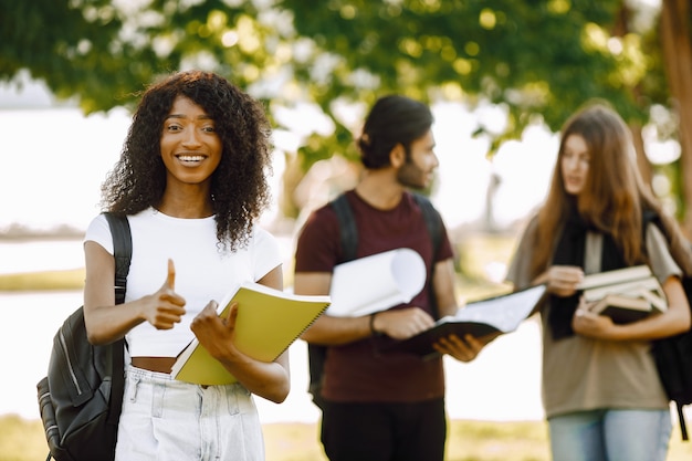 Focus on a african girl who standing sepately and shows thumb up. Group of international students standing together in park at university