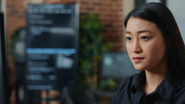 Focused asian software developer writing programming code and smiling sitting at desk in database development agency. Friendly coder computing big data casually in it startup office.