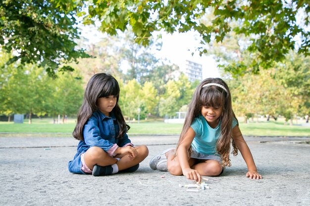 Focused black haired girls sitting and drawing on asphalt with colorful pieces of chalks. Front view. Childhood and creativity concept