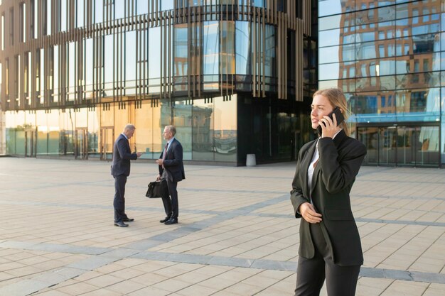 Focused businesswoman wearing office suit, talking on mobile phone outdoors. Businesspeople and city building glass facade in background. Copy space. Business communication concept